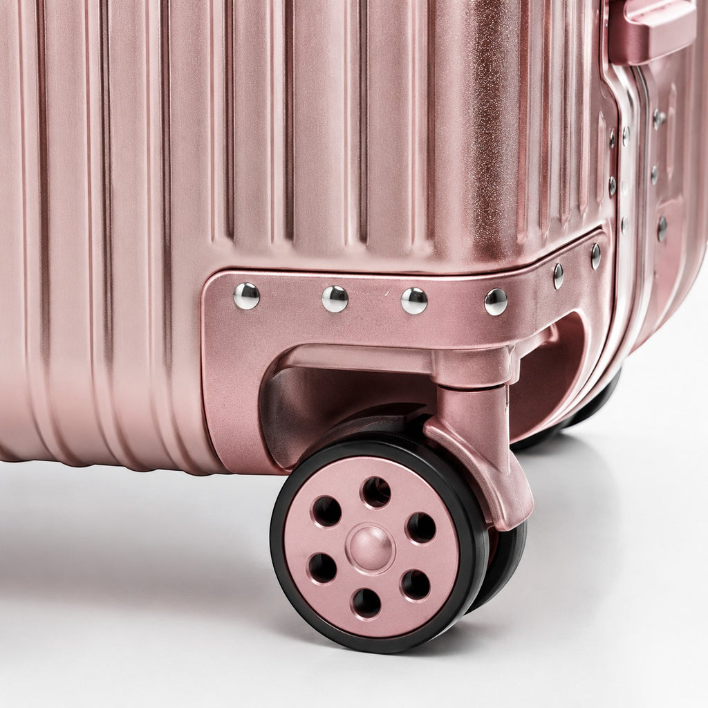 CASE.01 aluminum suitcase in rose gold with ribbed texture and black wheels. Features studded corner accents.